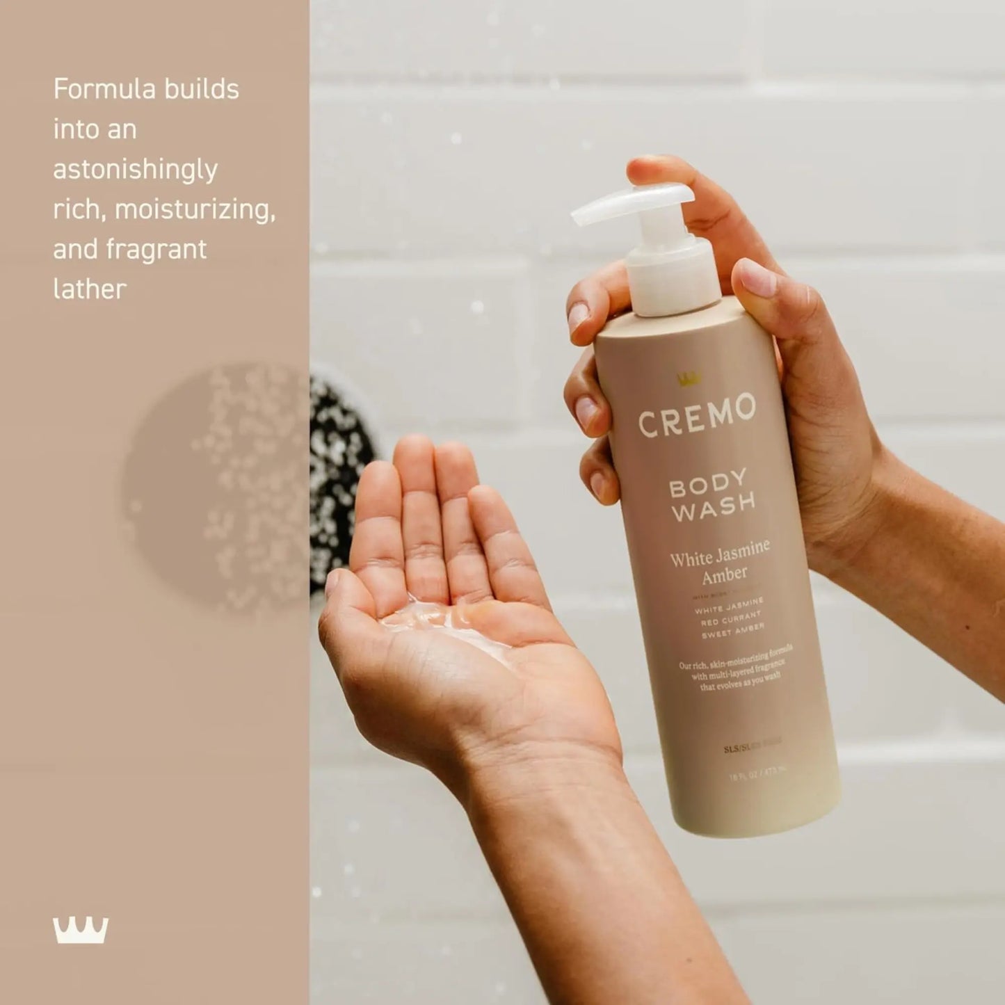 Cremo body wash bottle with a hand holding it and another hand with soap lather on a bathroom background.